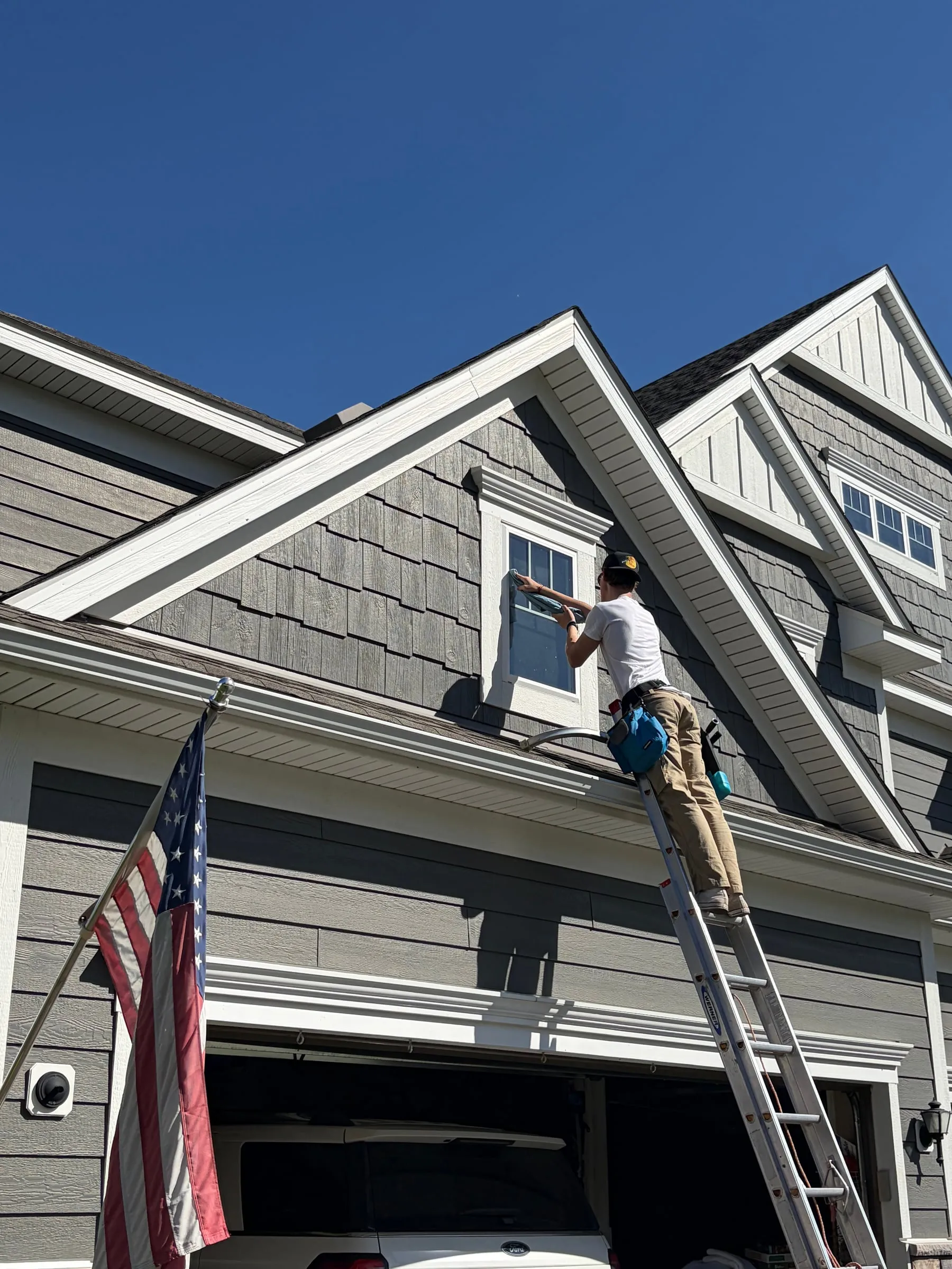 Top Haus crew member on a ladder cleaning a gable window, an American flag flying beside the garage